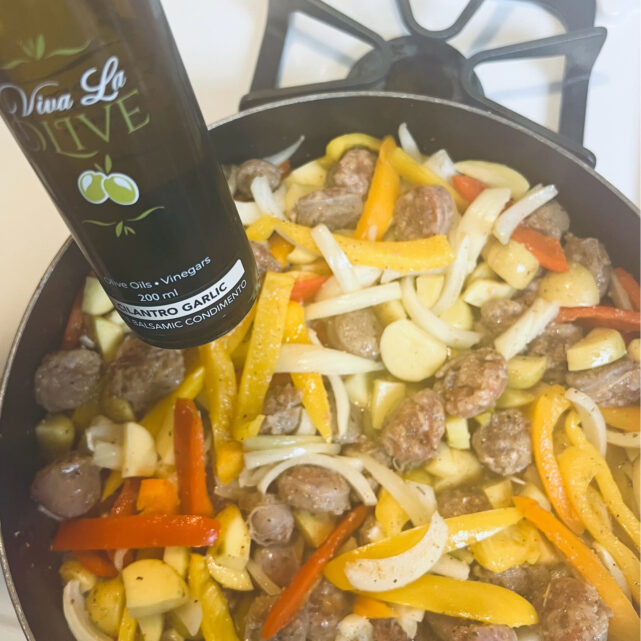 A skillet on a stove filled with sausage, peppers, and potatoes. A bottle of Cilantro Garlic White Balsamic Vinegar is nearby.